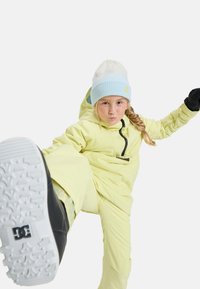 Gele skikleding met een capuchon en een ritszak, gecombineerd met een lichtblauwe gebreide muts. Zwarte sneeuwlaarzen met een witte zool.