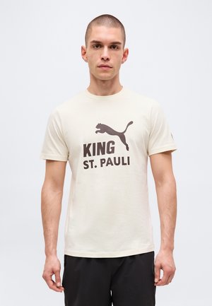 Jeune homme aux cheveux courts portant un T-shirt crème avec un logo Puma foncé et le texte "KING ST. PAULI", debout devant un fond uni.