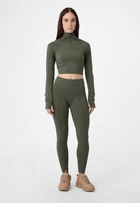 Oliven grøn atletisk sæt inkluderer en kort top med høj krave og lange ærmer, parret med tætsiddende leggings og brune atletik sko.