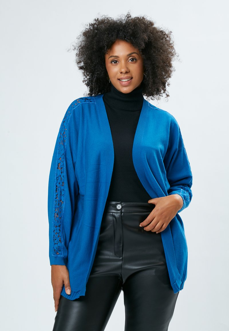 Hæklet blå cardigan med blondeaccentueret på ærmerne, åben frontdesign og ribbede manchetter, parret med en sort turtleneck og læderbukser.