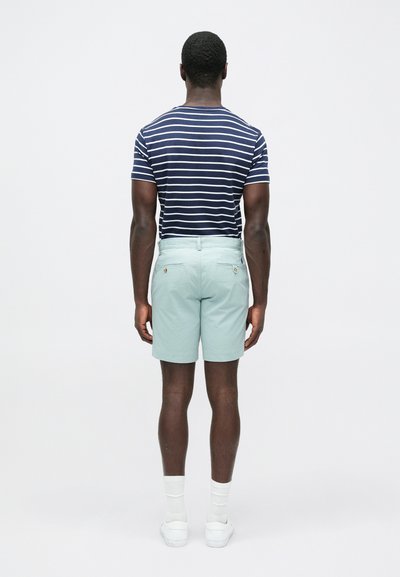 Μπλούζα κοντομάνικη με ρίγες σε ναυτικό μπλε και άσπρο, συνδυασμένη με ανοιχτό πράσινο σορτς· το μοντέλο εμφανίζεται από πίσω με άσπρες κάλτσες και παπούτσια.