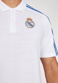 Polo blanc avec des rayures bleues sur les épaules et le logo brodé du club de football Real Madrid sur la poitrine, porté par une personne.