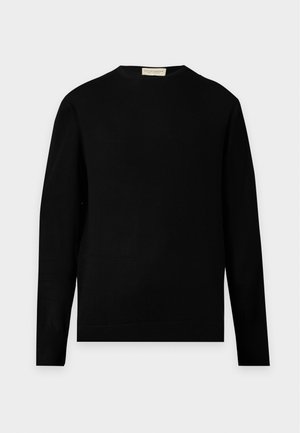 Mann trägt ein schwarzes Langarmhemd, schwarze weite Hosen und weiße Sneakers und steht vor einem schlichten hellen Hintergrund.