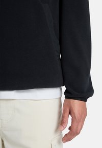 Sudadera de forro polar negra con un corte holgado, que presenta un bolsillo frontal, tejido texturizado y forro interior en contraste. Camisa blanca debajo.