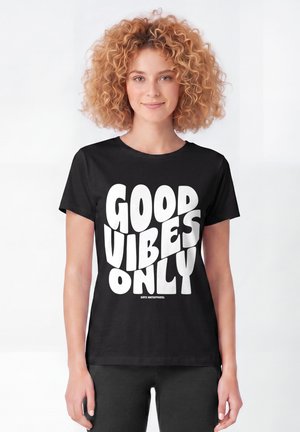 Jeune femme aux cheveux bouclés portant un t-shirt noir avec un grand texte blanc indiquant "GOOD VIBES ONLY" sur un fond blanc uni.