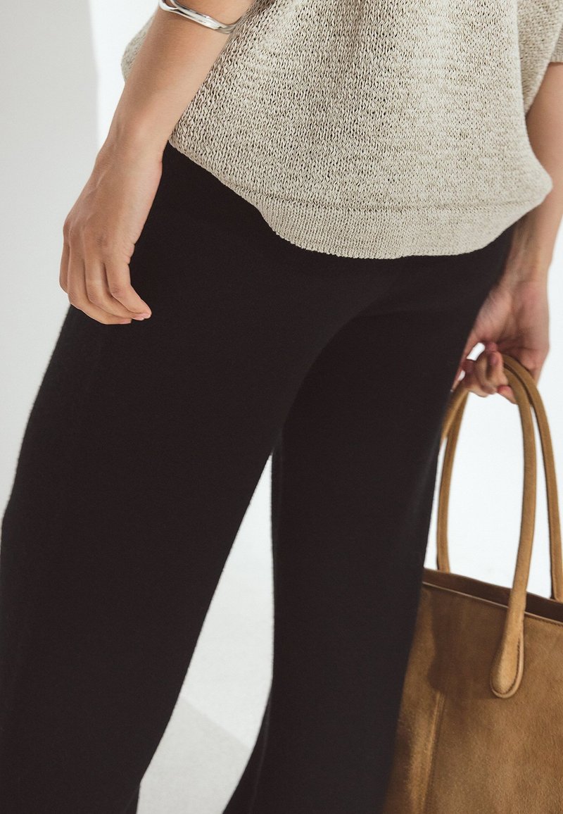 Personne portant un haut en maille beige et un pantalon noir, tenant un sac à main en daim marron par les anses, vue centrée sur le bas du torse et les mains.