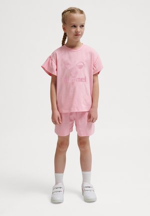Ung pige med fletninger iført pink t-shirt og shorts, hvide sokker og hvide sneakers, står mod en ensfarvet baggrund.