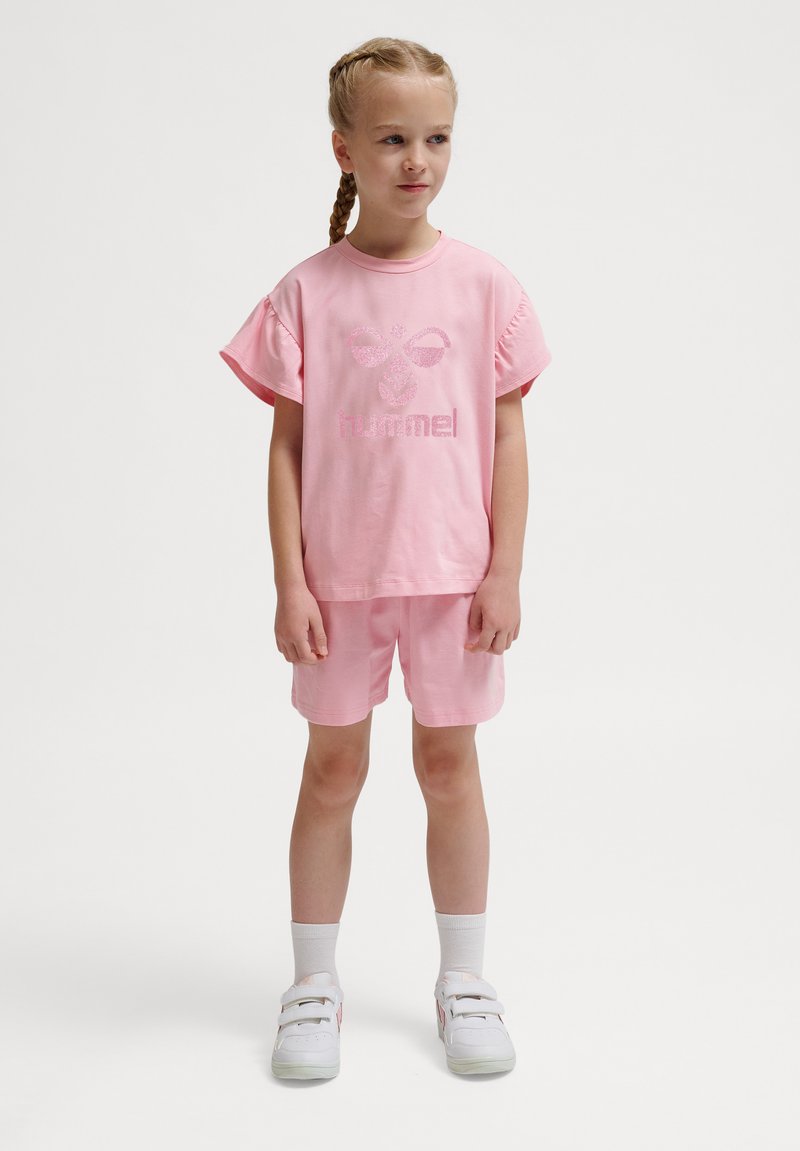 Ung pige med fletninger iført pink t-shirt og shorts, hvide sokker og hvide sneakers, står mod en ensfarvet baggrund.