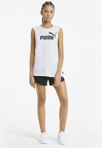 Vit ärmlös topp med svart "PUMA"-logotyp, kombinerad med svarta shorts. Modellen bär vita sneakers. Enkelt design, jämn textur.