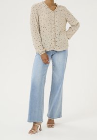 Blouse beige à pois noirs, col à volants et boutonnée à l'avant. Associée à un jean large bleu clair et des sandales à talons nude.