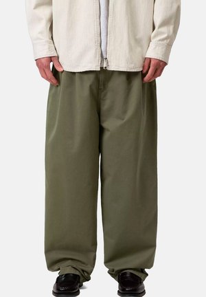Persona che indossa pantaloni larghi verde oliva, scarpe nere e una giacca beige con zip sopra una camicia bianca, in piedi davanti a uno sfondo uniforme.