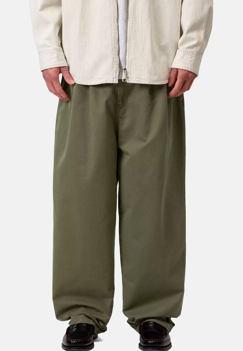 Persona con pantalones anchos color verde oliva, zapatos negros y una chaqueta beige con cremallera sobre una camiseta blanca, de pie frente a un fondo liso.