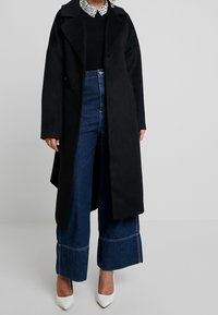 Lång svart kappa i mjukt material, med en skördkantad krage. Tillsammans med vida blå denimjeans, fållade med en stickad kant.