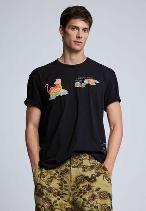 Joven con camiseta negra con diseños coloridos de leopardos y nubes, y pantalones amarillos con estampado floral, con las manos en los bolsillos.