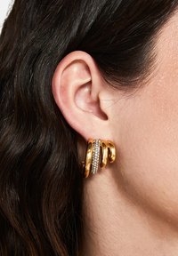 Guldtonade hoopsringar med tre sammanflätade band, varav ett är prytt med klara rhinestones och visar en slät, glänsande yta.