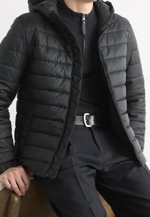 Personne portant une veste matelassée noire par-dessus un pull noir zippé, un pantalon noir et une ceinture noire, assise sur une chaise marron.