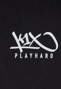 Sort stof med et hvidt grafisk design, der indeholder teksten "k1x" og "PLAYHARD" i en stiliseret skrifttype. Baggrunden er ensfarvet.