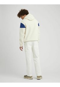 Un sweat à capuche blanc avec des accents bleu marine sur les épaules, associé à un pantalon cargo blanc. Le sweat présente une capuche avec cordon de serrage et une texture douce.