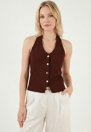 Blondínka v tmavohnedom bezrukávovom halter top s gombíkmi a svetlo béžových nohaviciach, jedna ruka v vrecku na jednofarebnom pozadí.