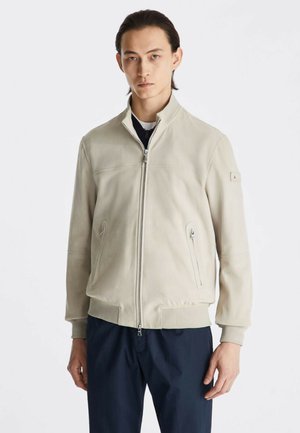 Mann mit glatten dunklen Haaren trägt eine hellbeige Zip-up-Jacke und dunkle Hose und steht vor weißem Hintergrund.