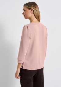 Frau mit glattem, blondem Haar, die seitlich schaut, trägt eine hellpinkfarbene Bluse mit langen Ärmeln und schwarze Hosen vor einem einfarbigen weißen Hintergrund.