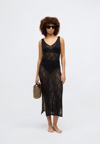 Μαύρο πλέγμα κάλυψης με γεωμετρικό σχέδιο, χωρίς μανίκια, πλαϊνές σχισμές και μήκος μέχρι το μισό μοσχάρι. Συνδυασμένο με μια ριγέ τσάντα.