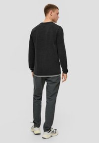 Schwarzer strukturierter Pullover mit langen Ärmeln, rundem Ausschnitt und grauen Hosen. Die Schuhe bestehen aus einer Mischung aus Schwarz und Beige mit markanten Designelementen.