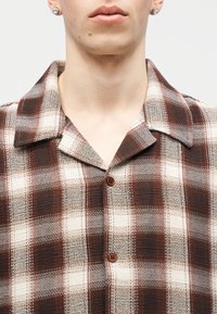 Νεαρό άτομο που φοράει καφέ-κρεμ πουκάμισο με καρό και ευρύ γιακά, καθώς και ασημένια σκουλαρίκια με στρας.