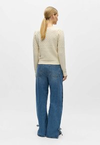 Femme avec une queue de cheval blonde portant un pull en tricot crème et un jean bleu ample avec des liens aux ourlets, debout de dos sur un fond blanc.
