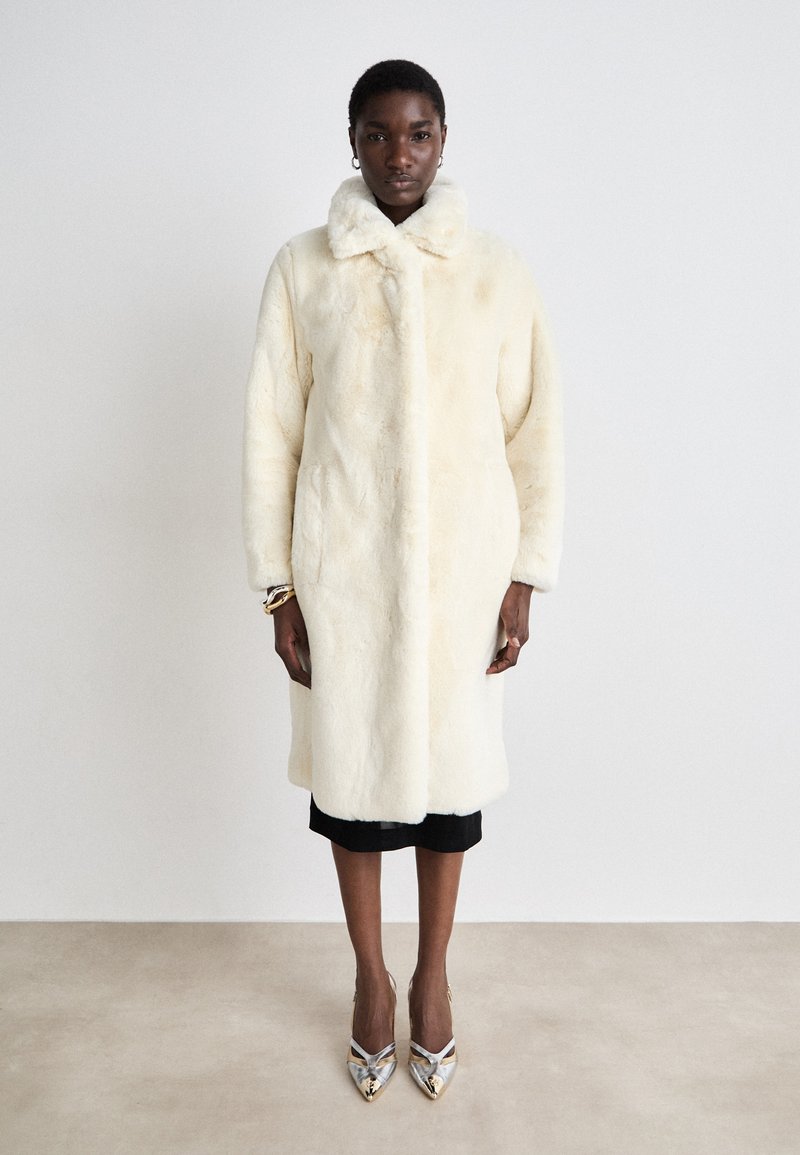 Lang creme faux fur frakke med høj krave, sidelommer og afslappet pasform, parret med sølv højhælede sko. Glat tekstur.