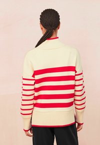 Person med fletning bærer en cremet sweater med horisontale røde striber, står mod en ensfarvet lys baggrund, set fra baghagen.