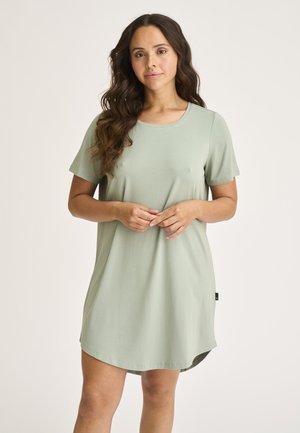 Kvinde med langt bølget hår iført en løs, lys grøn kortærmet kjole, stående med hænderne foldet foran sig mod en ensfarvet baggrund.