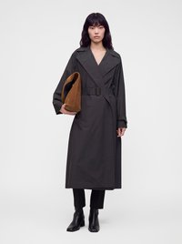 Femme aux cheveux noirs et raides porte un long trench-coat noir ceinturé, un pantalon noir, des bottines noires, tenant un grand sac en daim marron.