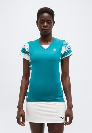 Žena nosi čistomodrý športový top s krátkymi rukávmi s bielymi vzorovanými rukávmi a bielu športovú sukňu s čiernym logom na leme.