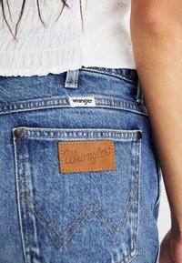 Mėlyni džinsai su odiniu Wrangler logotipu ant kišenės, kontrastingais siuvinėjimais ir klasikiniu penkių kišenių dizainu.