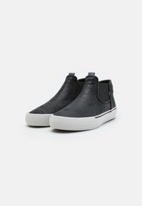 Zapatos negros tipo slip-on con una parte superior texturizada, paneles laterales elásticos, suela de goma blanca y una tira en el talón para ponerlos y quitarlos fácilmente.