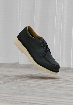 Scarpa nera in pelle con lacci e suola beige che fluttua sopra un pavimento in legno e tende bianche sullo sfondo.