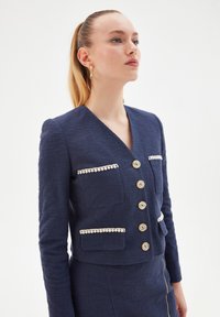 Navy teksturoitu takki syvällä V-kaula-aukolla, jossa on kerma-väriset reunat taskuissa ja viisi pyöreää nappia. Slim fit ja pitkät hihat.