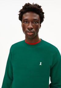 Felpa verde con colletto a girocollo, tessuto testurizzato, logo bianco sul petto, indossata con una camicia rossa sotto, modello con capelli corti e ricci.