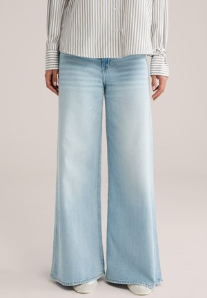 Žena oblečená v svetlomodrých širokých džínsach a béžovo-bielej vertikálne pruhovanej košeli na gombíky, stojaca pred neutrálnym pozadím.