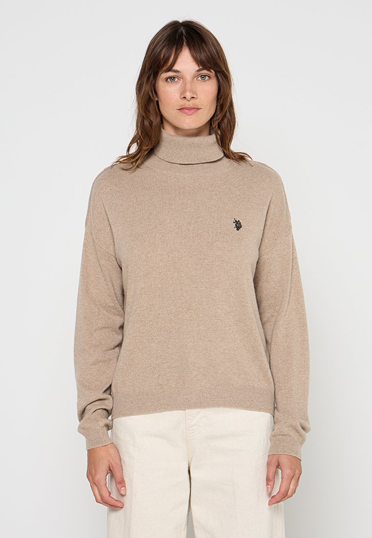 Maglione beige a collo alto realizzato in materiale morbido, con maniche lunghe e un piccolo logo nero sul lato sinistro del petto.