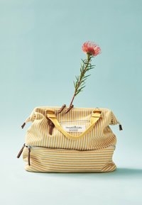 Bolsa de tela amarilla a rayas con acentos y cremalleras en marrón. De la apertura de la bolsa brota una flor, añadiendo un toque decorativo.