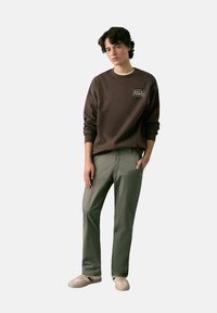 Felpa marrone con scollo rotondo con una toppa logo, abbinata a pantaloni verde chiaro e sneakers beige basse con dettaglio verde.