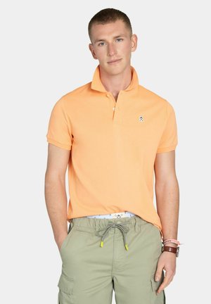 Joven con una camisa polo de manga corta color melocotón metida dentro de unos pantalones cortos cargo verde oliva con cordón, de pie con las manos en los bolsillos.