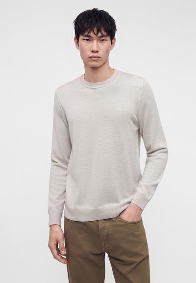 Jeune homme aux cheveux noirs portant un pull beige clair et un pantalon marron, debout avec une main dans la poche sur un fond uni.