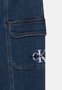 Ciemnoniebieski jeans z pomarańczowym przeszyciem, z płaską kieszenią i wszytym logo "Calvin Klein" w białej i czarnej nitce.