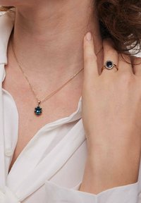 Złoty naszyjnik z kwiatowym wisiorkiem z niebieskimi kamieniami. Ponadto złoty pierścionek z ciemnym kamieniem w centrum otoczonym mniejszymi kamieniami.