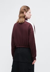 Bordo pulóver s kapucňou a vysokým golierom, elastickým pásom a kontrastnými bielymi bočnými pruhmi. Mäkke materiál, voľný strih a dlhé rukávy.