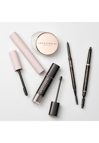 Fünf Kosmetikprodukte auf einem weißen Hintergrund angeordnet: ein Glasbehälter mit roségoldenem Deckel, zwei Augenbrauenstifte, eine Wimperntusche und ein kleines Röhrchen.