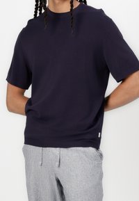 Pull en maille bleu marine à manches courtes avec un col rond et un ourlet côtelé, associé à un pantalon rayé gris clair. Design simple, sans ornements.